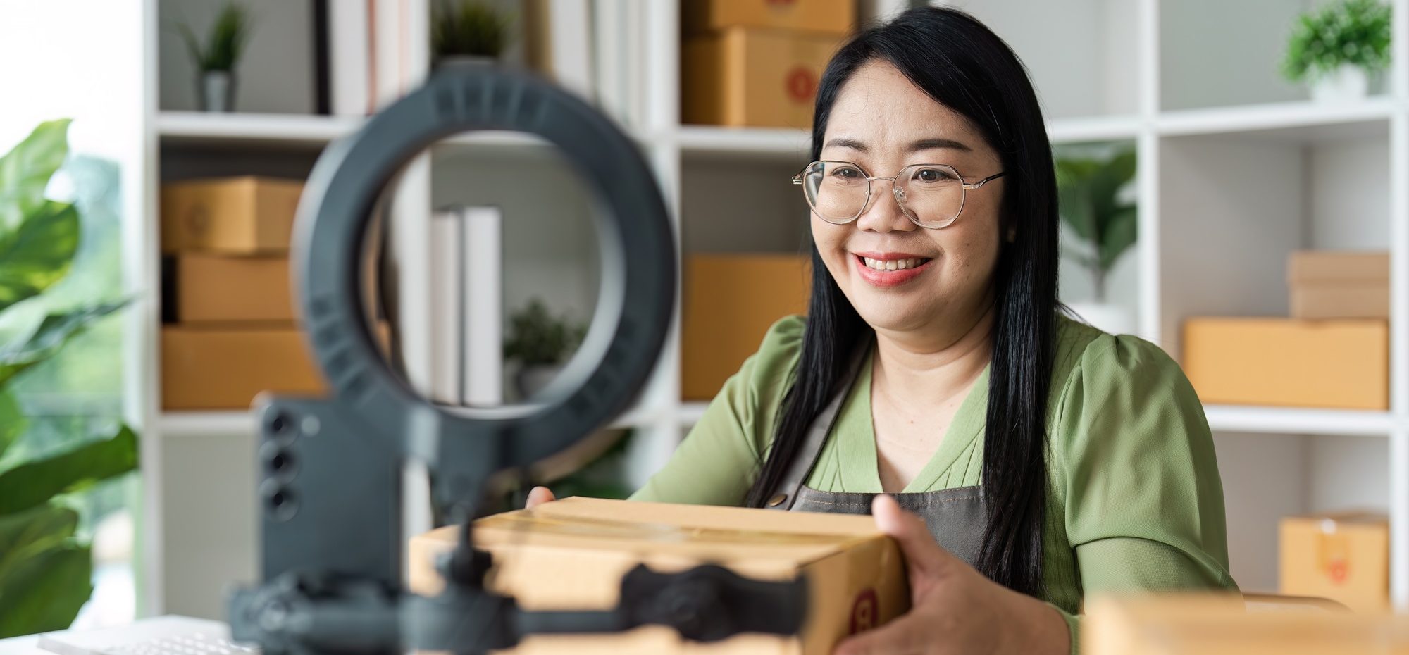 Smiling Woman Conducting Live Selling Online with Packaging Boxes in Modern Home Office Setup